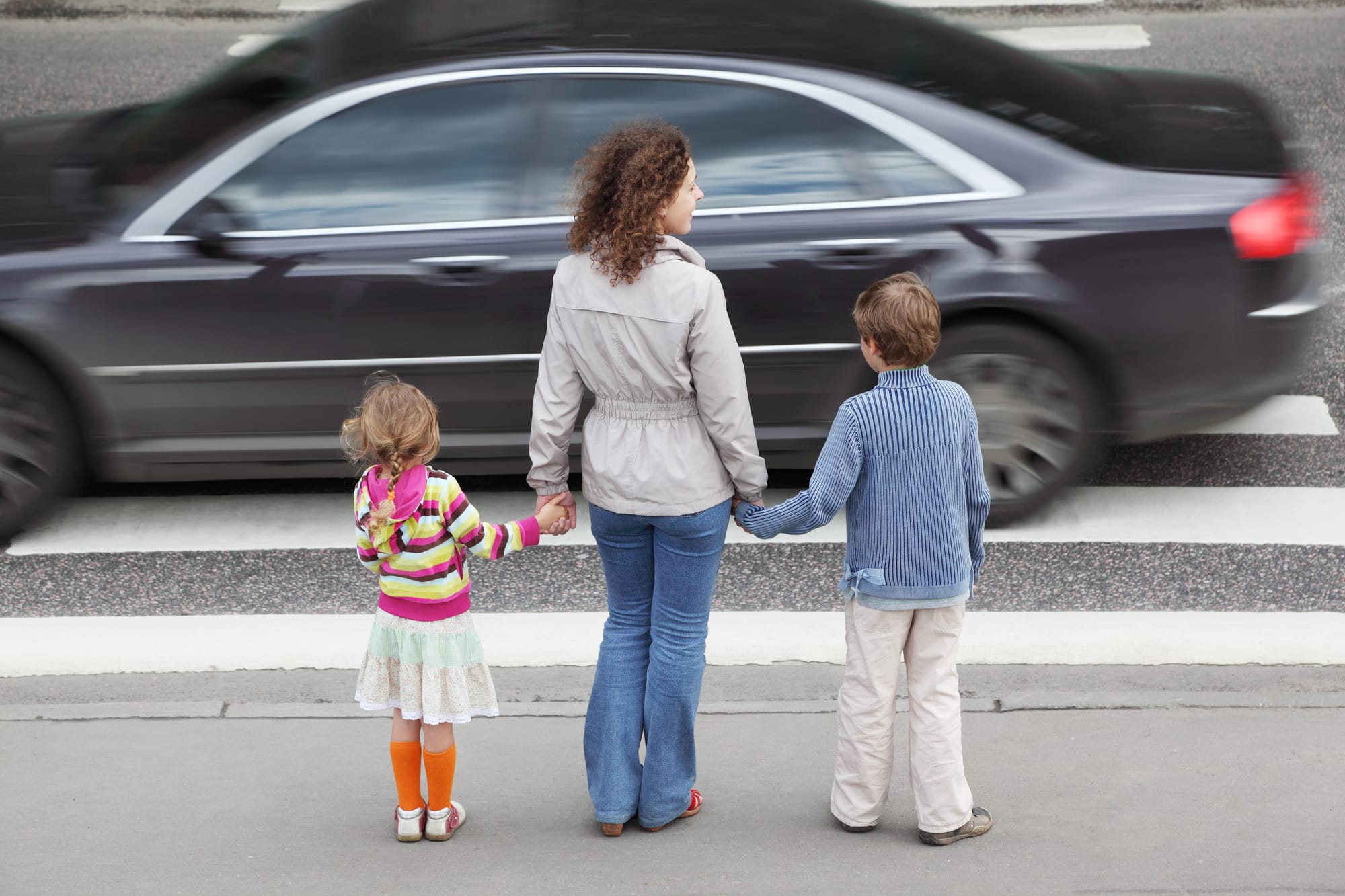 A mother holds hand of young daughter and son and standing near crosswalk as a black car speeds by them.