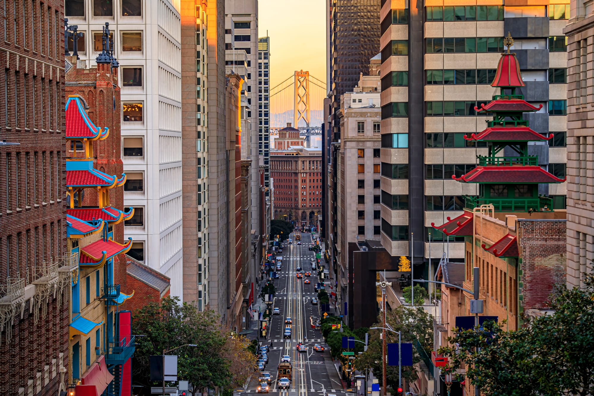 View of California Street near China Town and the Financial District, with Chinese pagoda towers and the Bay Bridge at sunset with cars on the street.