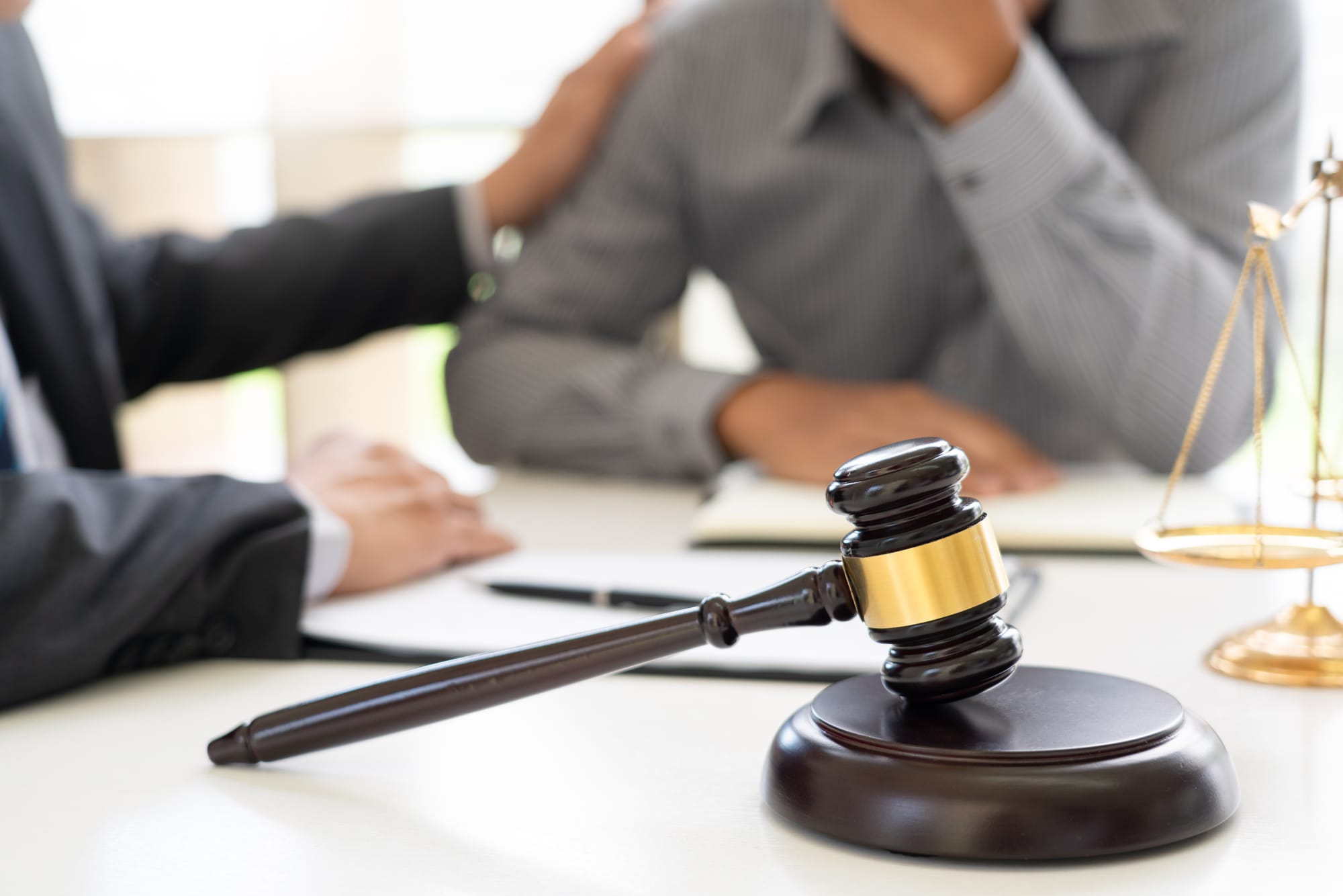 A blurred image of a lawyer having meeting and consoling his male client with paperwork, gavel and scales of justice on the desk where they're meeting.