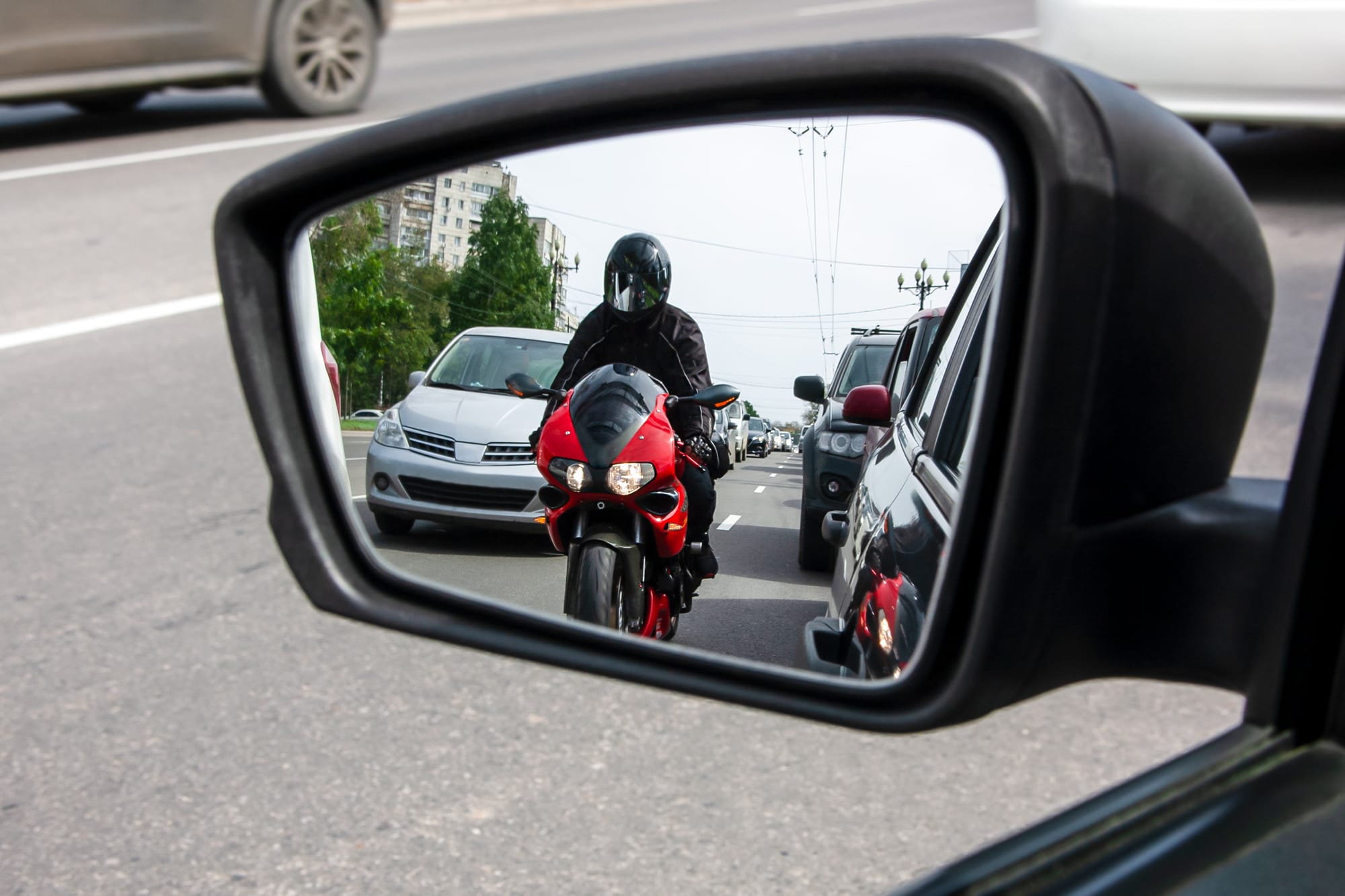 The side mirror of a car in traffic reflects a motorcyclist who is between cars.