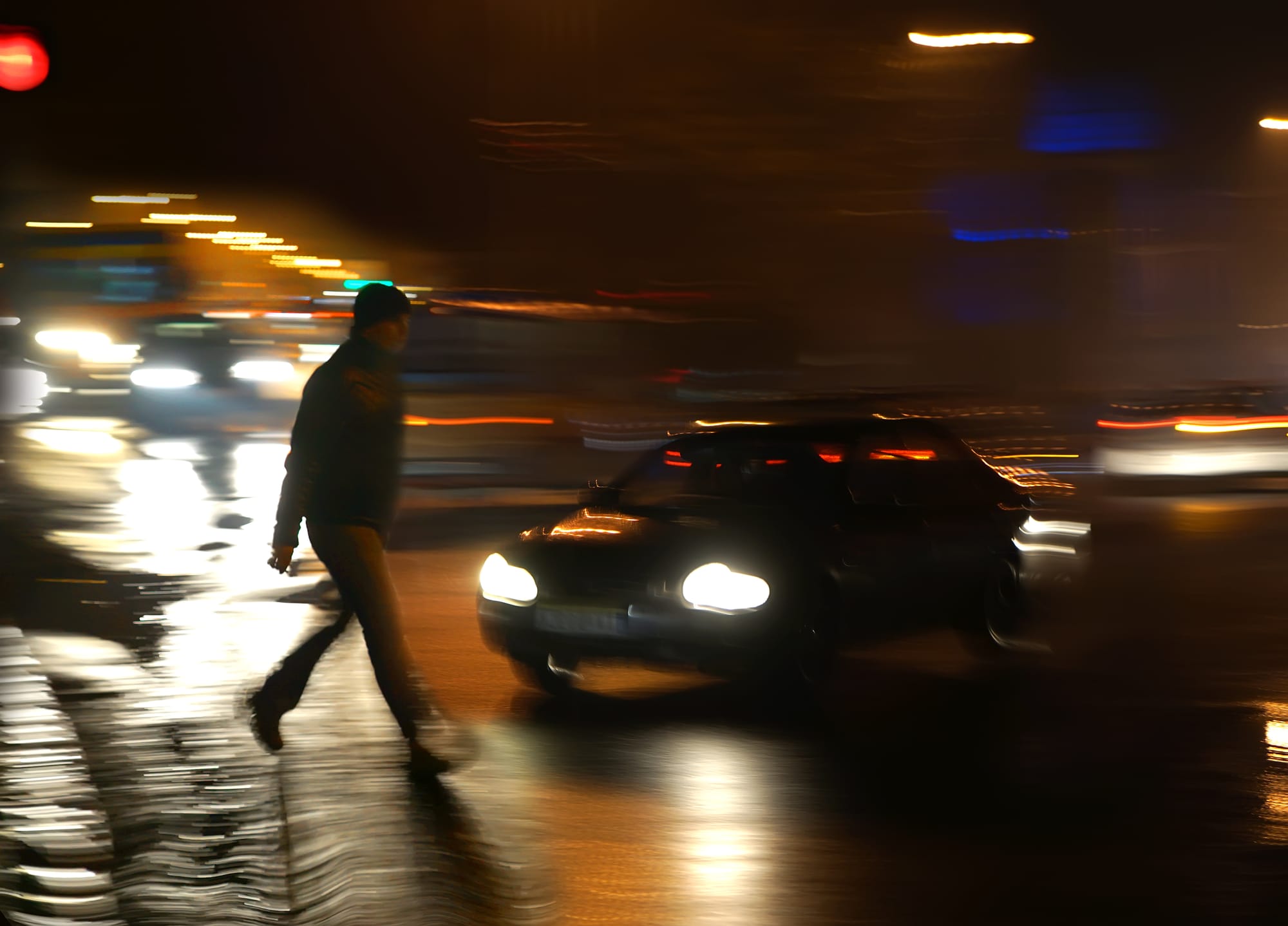 A male pedestrian crossing a street at night with a blurred car coming towards him.